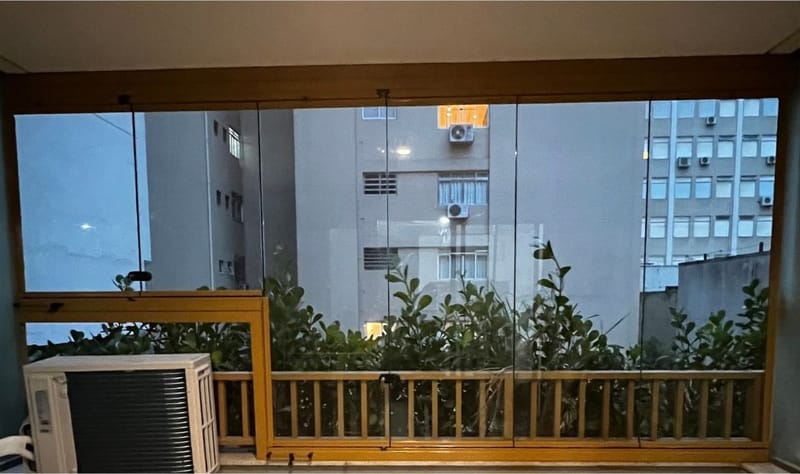 Sacada de apartamento fechada com envidraçamento de vidro e esquadria em tom amadeirado, vista de dentro ao entardecer. Obra da VitraAlu em Jardim Paulista, São Paulo.