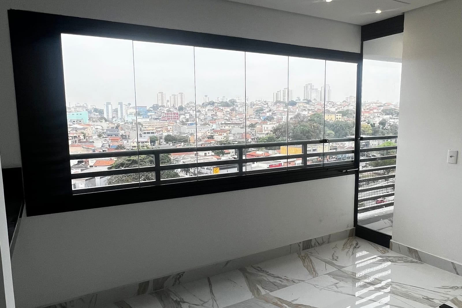 Sacada de apartamento envidraçada com esquadria de alumínio preta e piso porcelanato, com vista aberta para a cidade. Obra da VitraAlu em Casa Verde, São Paulo.